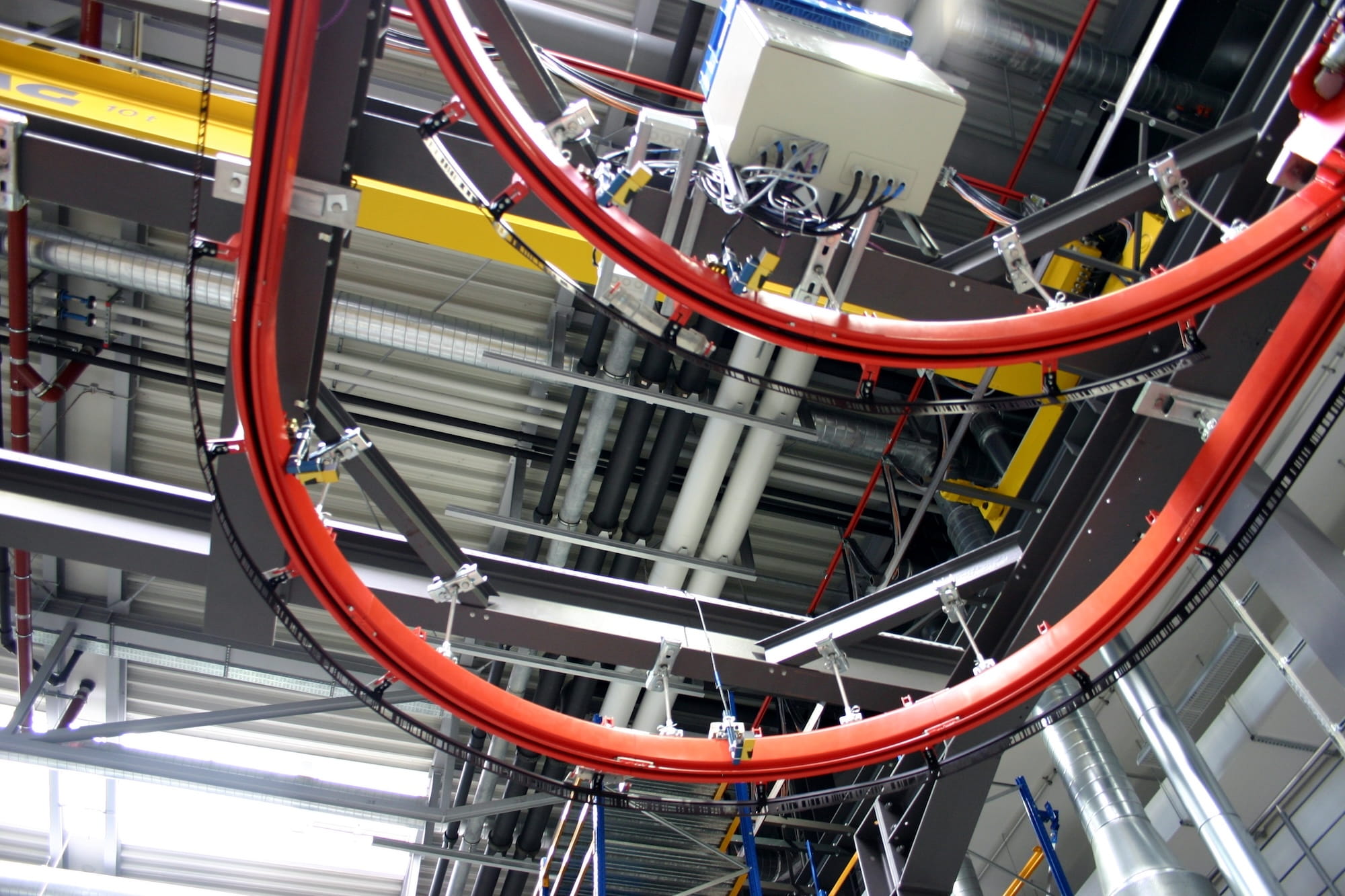 overhead view of automated red rail crane system with sensor-equipped hoists moving along curved tracks in an automotive manufacturing facility for intelligent material handling and part transport above the production floor